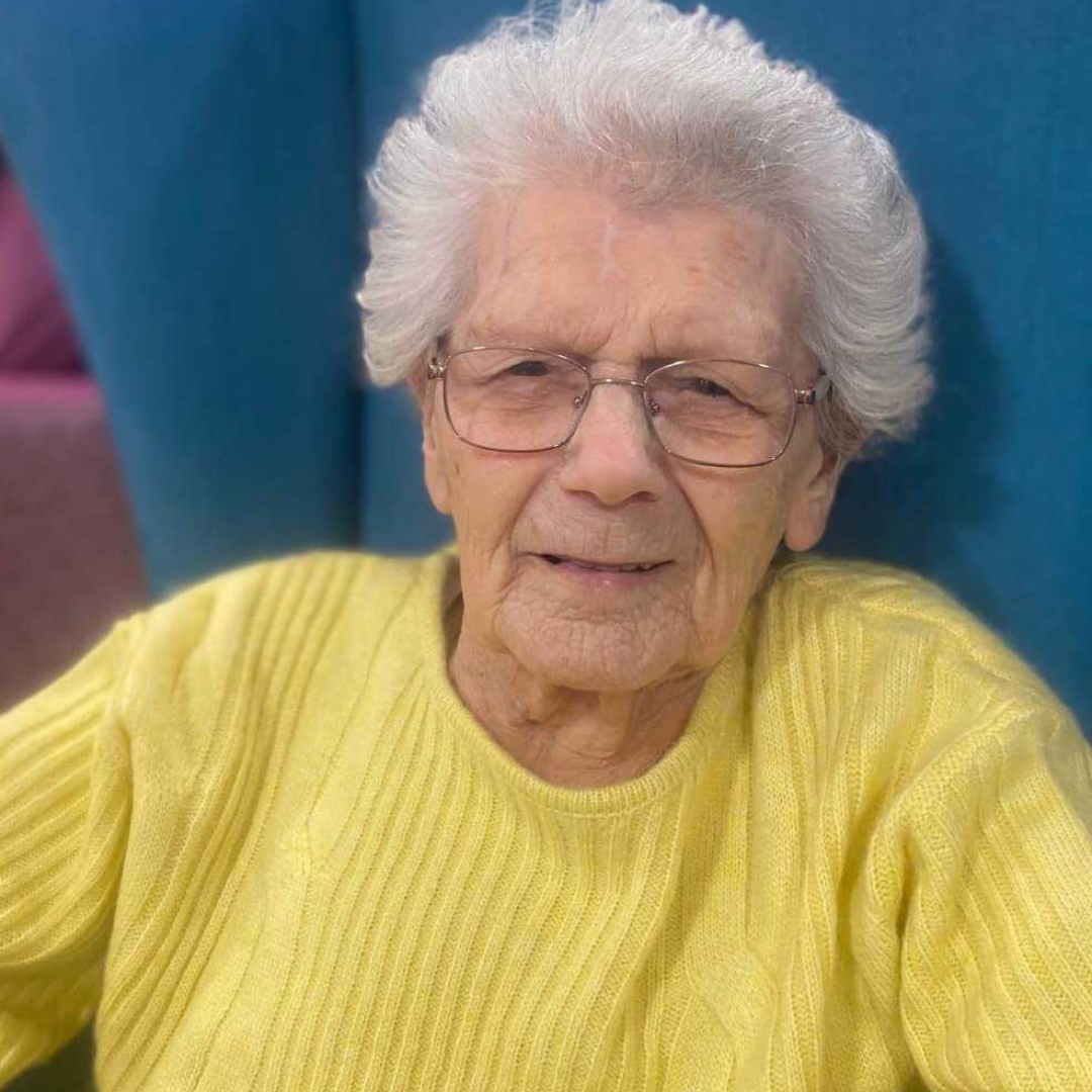 A woman in a yellow jumper sits comfortably in a blue wingback chair, as she celebrates her 102nd birthday
