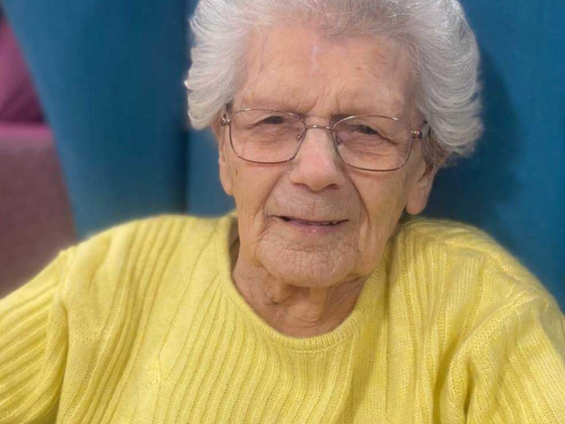 A woman in a yellow jumper sits comfortably in a blue wingback chair, as she celebrates her 102nd birthday