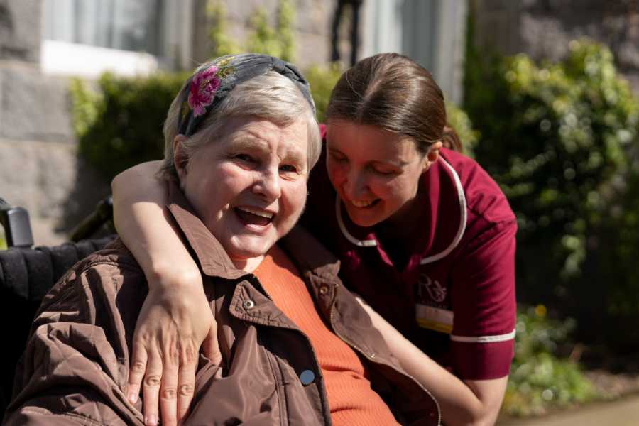 Caregiver offers a warm hug to a resident in a peaceful garden setting. The resident smiles, enjoying the moment of comfort and connection