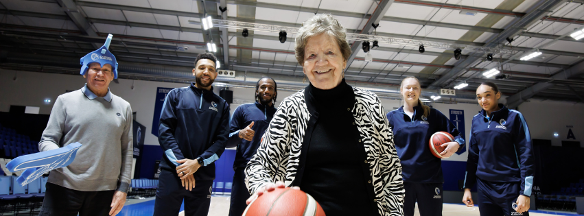 A care home resident at the forefront is bouncing a basketball, there are four Gladiator basketball players behind her and another care home resident with a foam finger and Gladiators headwear