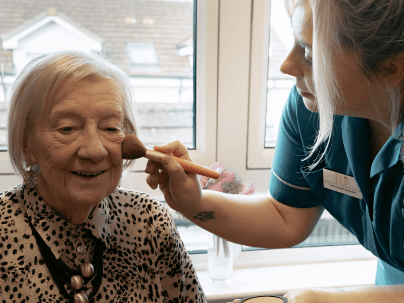 A caregiver in a teal uniform gently applies makeup to an elderly woman's cheek using a large soft brush. The woman, wearing a patterned blouse and a statement necklace, has a soft smile as she receives the pampering.