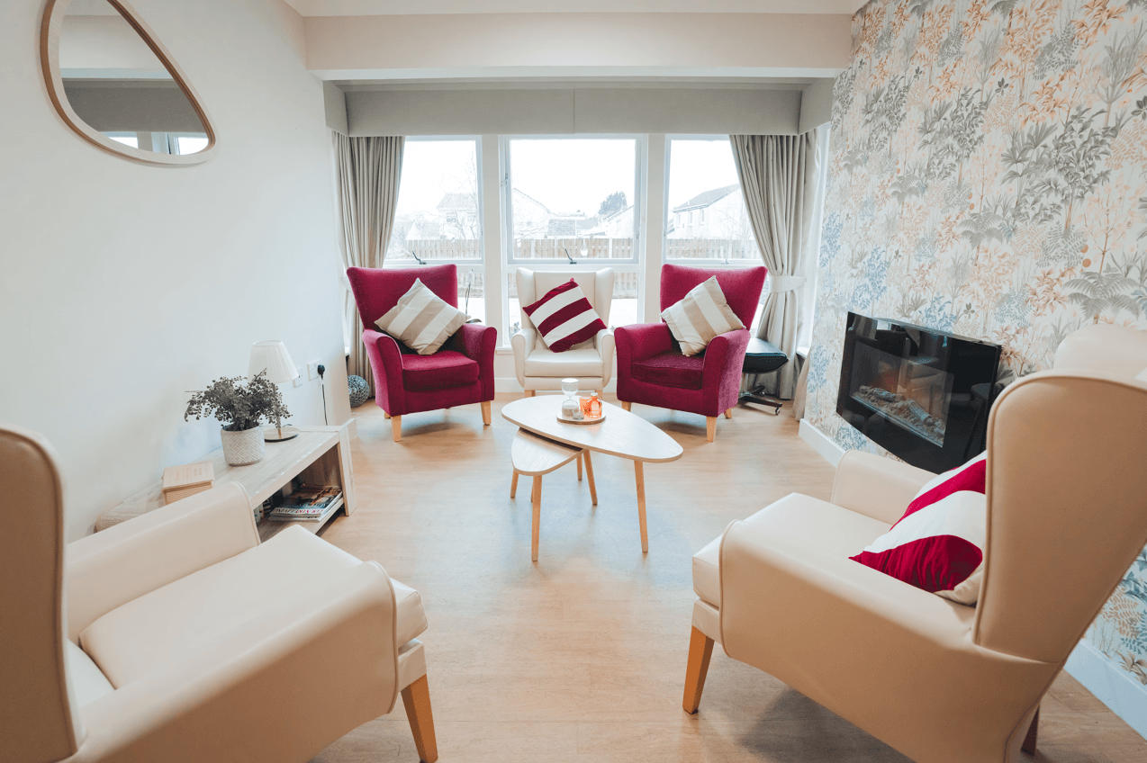 A sunlit lounge area designed for socialisation. Several plush armchairs in cream and deep pink are arranged around a modern tiered coffee table. The room features a floral accent wall with a built-in electric fireplace, light wood flooring, and large windows that provide a clear view of the outdoors.