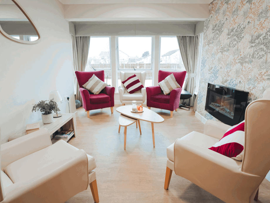 A sunlit lounge area designed for socialisation. Several plush armchairs in cream and deep pink are arranged around a modern tiered coffee table. The room features a floral accent wall with a built-in electric fireplace, light wood flooring, and large windows that provide a clear view of the outdoors.