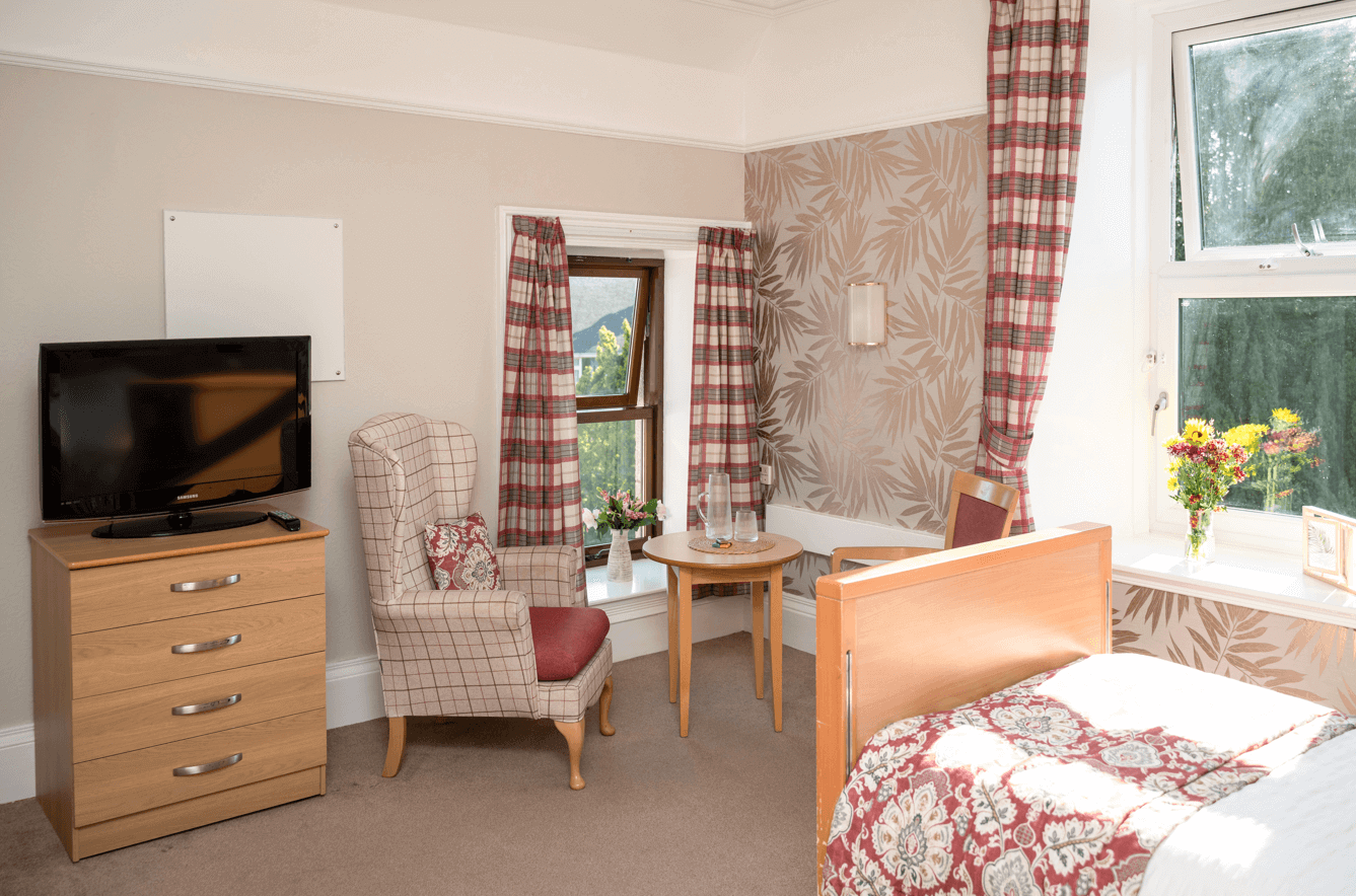 A warm and cosy copper and red bedroom features a comfortable corner seating area with bright windows and a TV on a set of drawers.