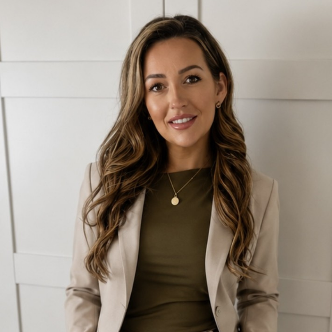 Roslyn, care home manager, stands wearing a nude blazer and high-necked blouse. She has long dark curled hair and stands in-front of a neutral background. 
