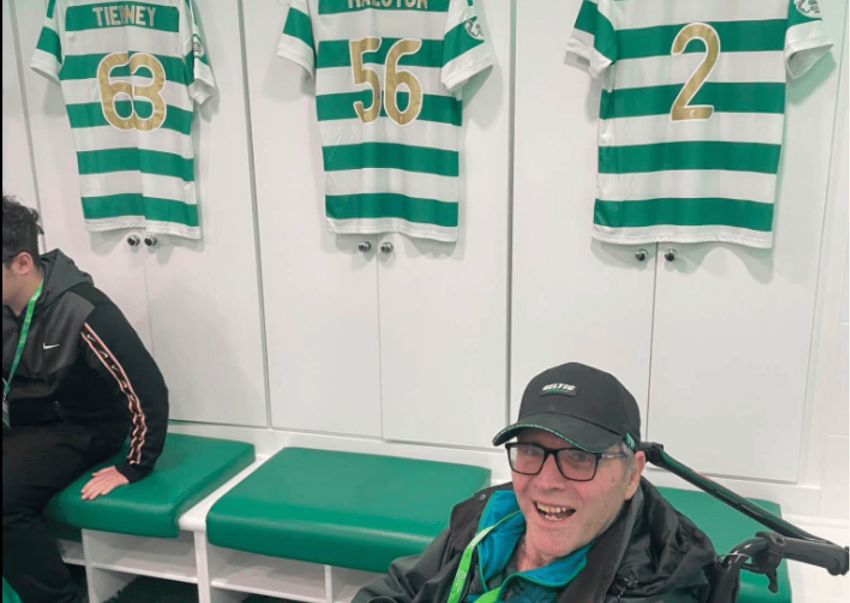 Three Celtic Player's Football kits hang on a white wall with green seat cushions and a long bench beneath. Care Home resident David is smiling in front of the shirts, he wears a black cap and glasses. 