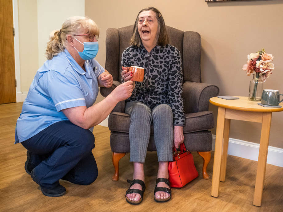 A staff member kneels down beside a resident enjoying a cup of tea in an armchair