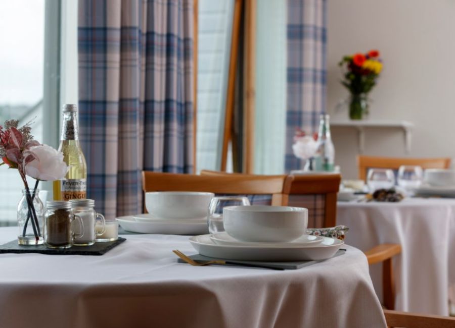 A dining room set up with a table and 4 chairs, napkins, plates, cutlery and flowers. There are windows with curtains to the rear 