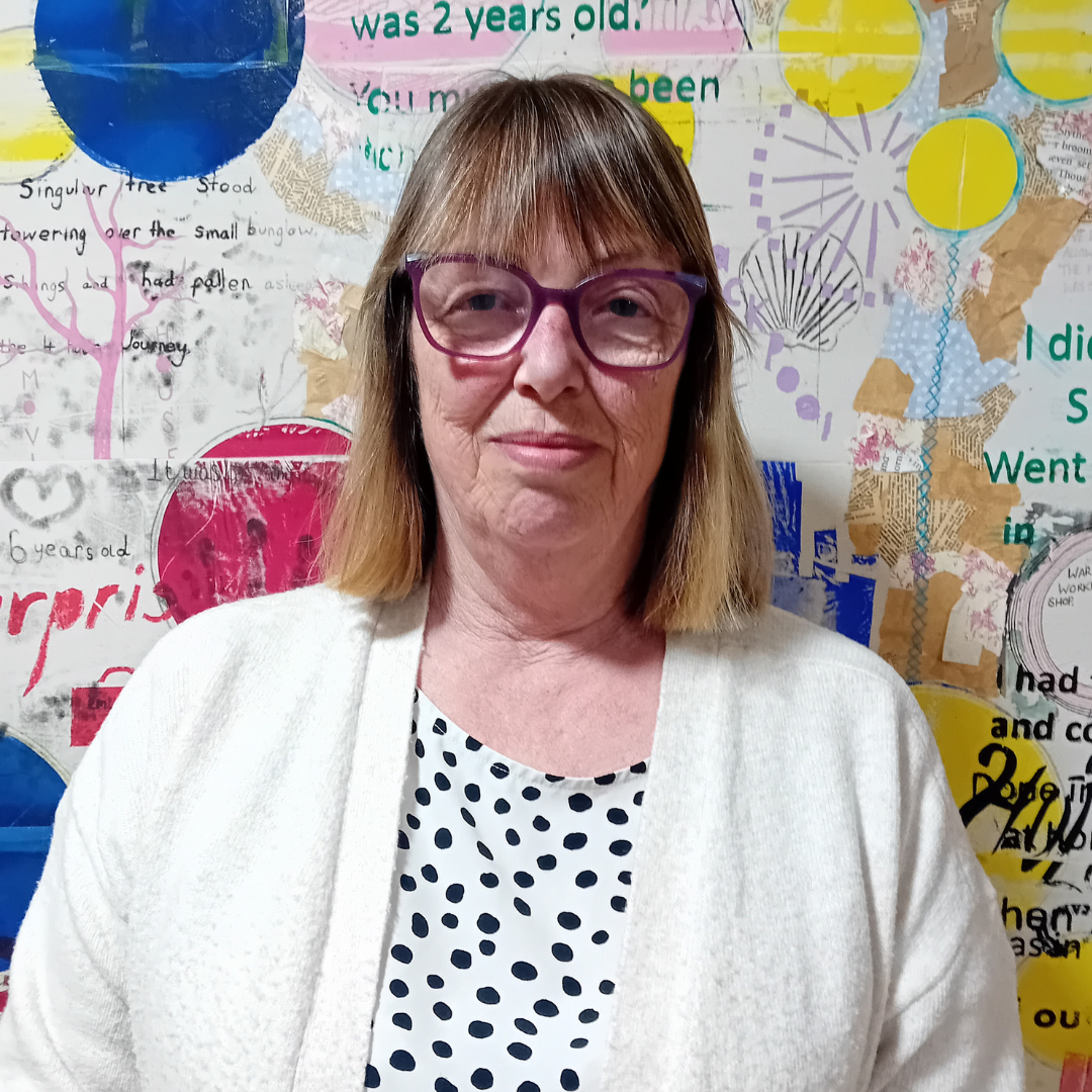 Care home manager Judith has shoulder length brown hair and glasses. She is wearing a white polka dot blouse and a white cardigan. 