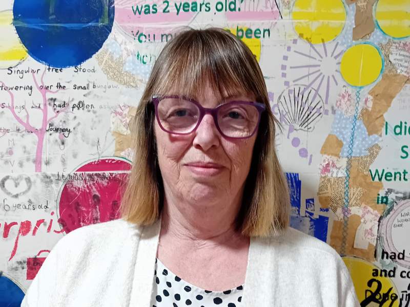 Care home manager Judith has shoulder length brown hair and glasses. She is wearing a white polka dot blouse and a white cardigan. 