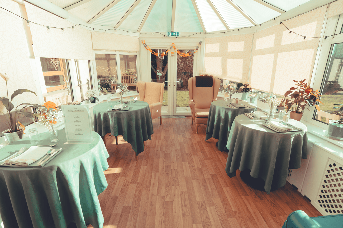 A wide view of a bright conservatory used as a dining area. Several round tables with green tablecloths are spaced out across the wood-laminate floor. Large windows and a glass ceiling allow for plenty of natural light.