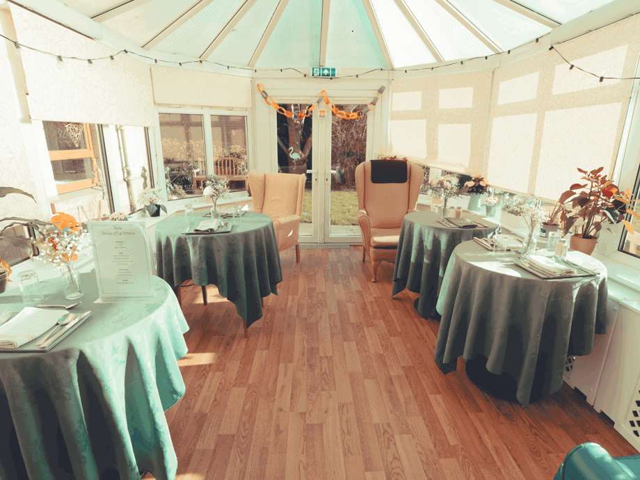 A wide view of a bright conservatory used as a dining area. Several round tables with green tablecloths are spaced out across the wood-laminate floor. Large windows and a glass ceiling allow for plenty of natural light.