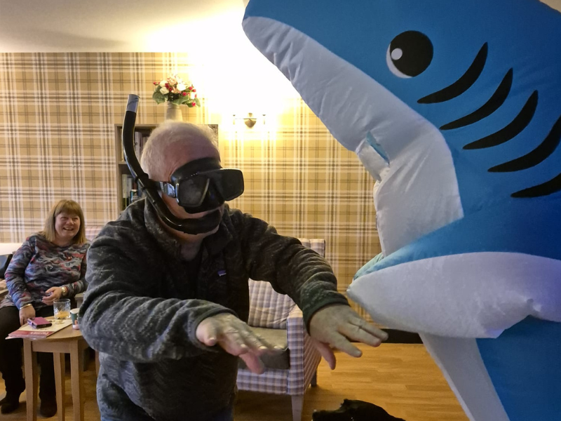A man wearing a snorkel mask and goggles interacts playfully with a large, bright blue inflatable shark costume. A black dog looks up at the man and the shark, while a seated woman in the background smiles. The scene suggests a playful, sea-themed event.