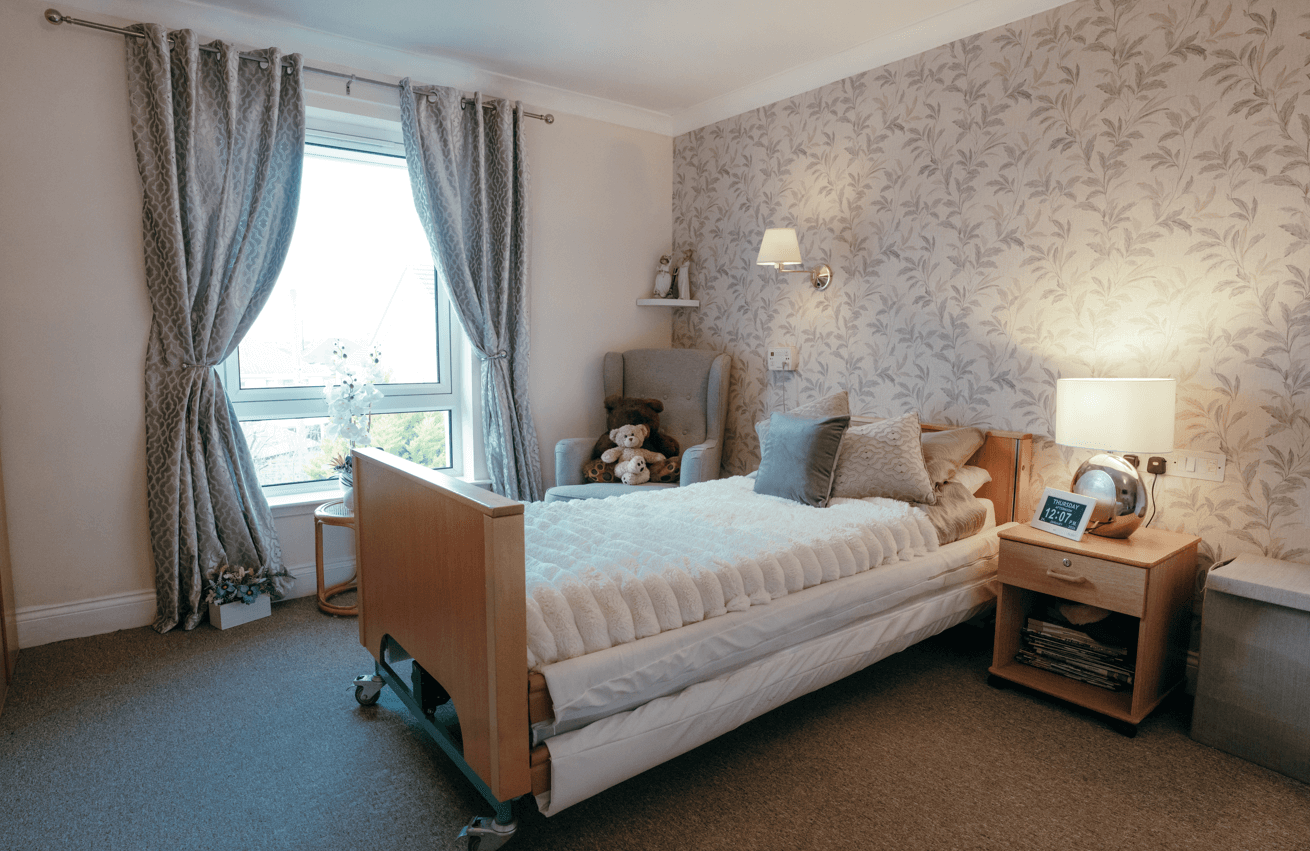 A well-lit bedroom featuring a single adjustable bed with white bedding. The room includes a patterned feature wall, a wooden bedside table with a lamp and digital clock, a grey armchair with teddy bears, and a large window with grey curtains.