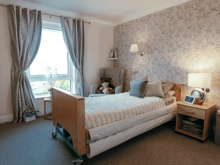 A well-lit bedroom featuring a single adjustable bed with white bedding. The room includes a patterned feature wall, a wooden bedside table with a lamp and digital clock, a grey armchair with teddy bears, and a large window with grey curtains.