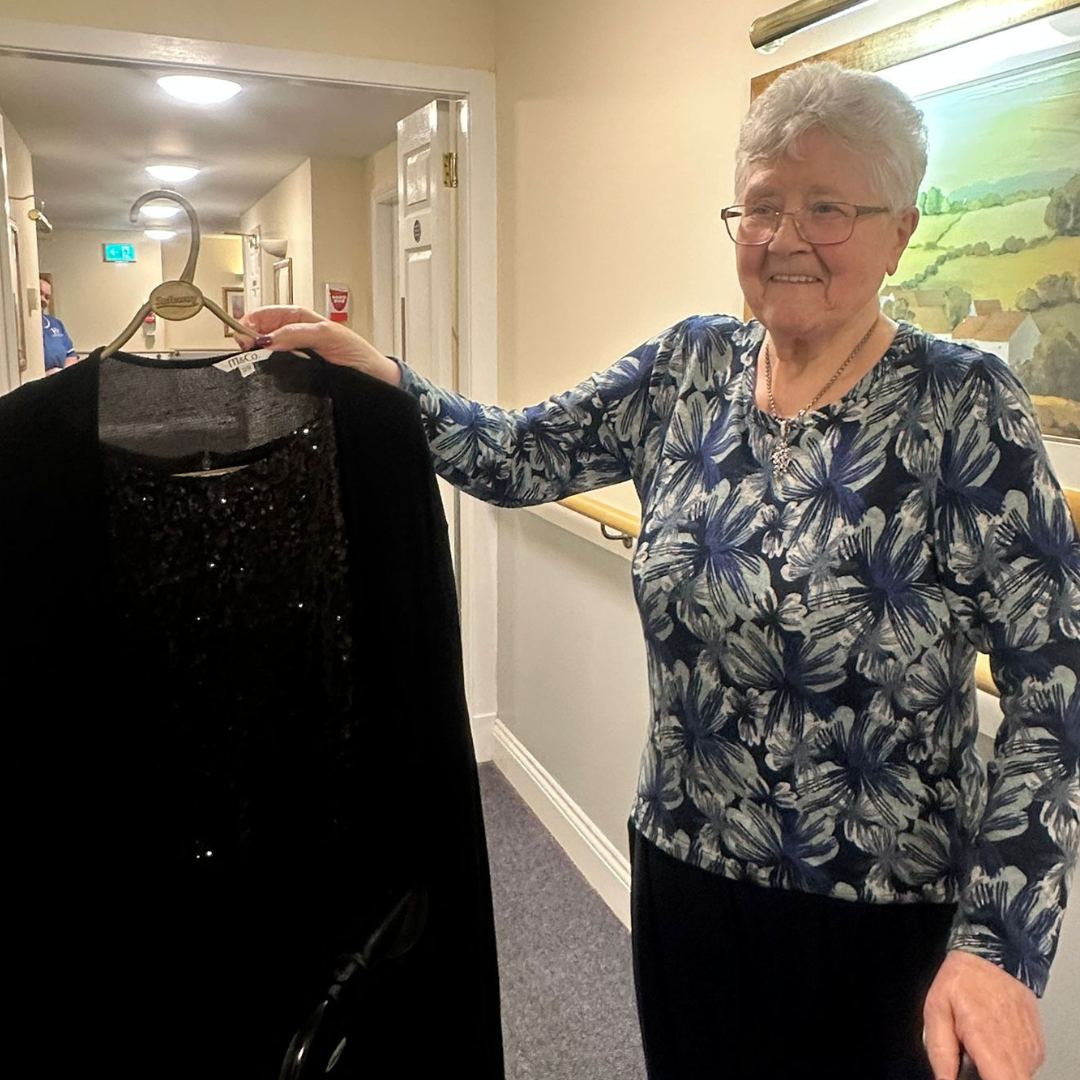 A woman with short hair and glasses holds up a black sparkly dress 
