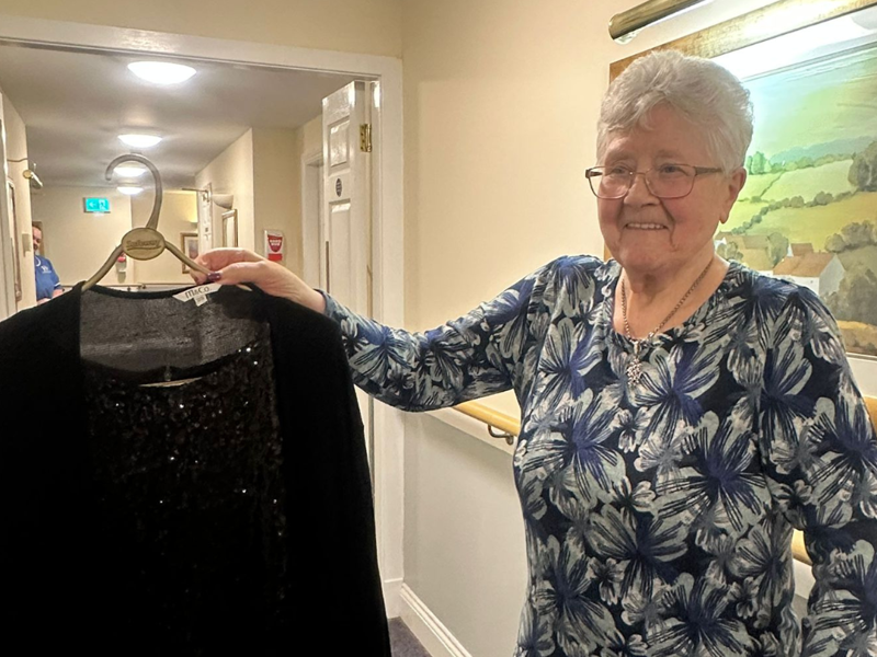 A woman with short hair and glasses holds up a black sparkly dress 