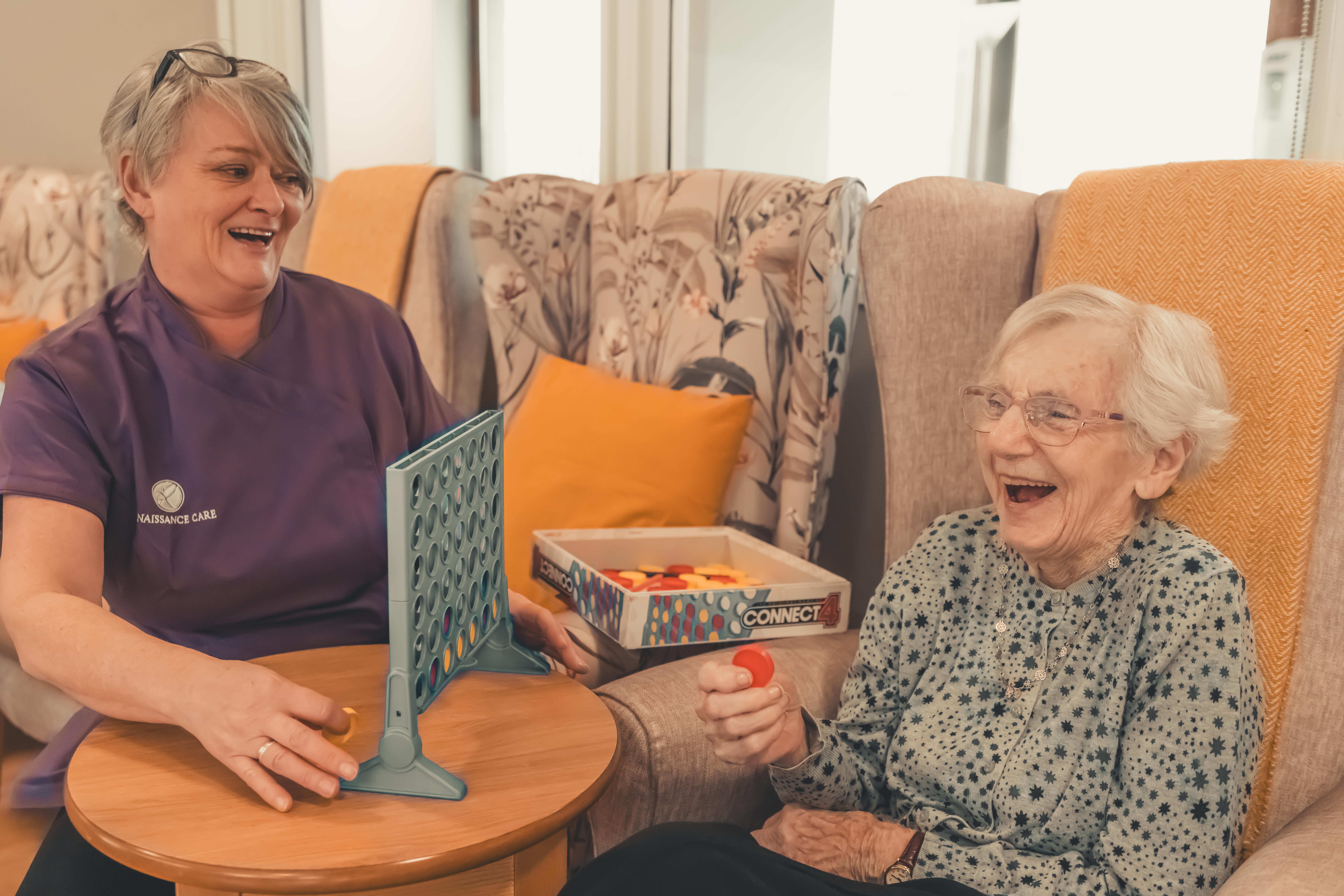A female resident and a caregiver in a purple uniform sit together in a lounge playing a game of Connect 4. Both are laughing heartily as the resident prepares to drop a red chip into the grid. The room is filled with comfortable, patterned wingback chairs.