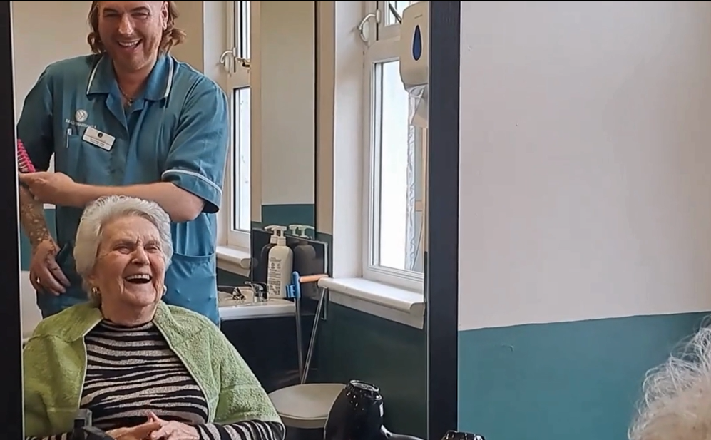 A resident sits in a hairdressers chair within the care home. She is laughing as the carer affectionately styles her hair. 