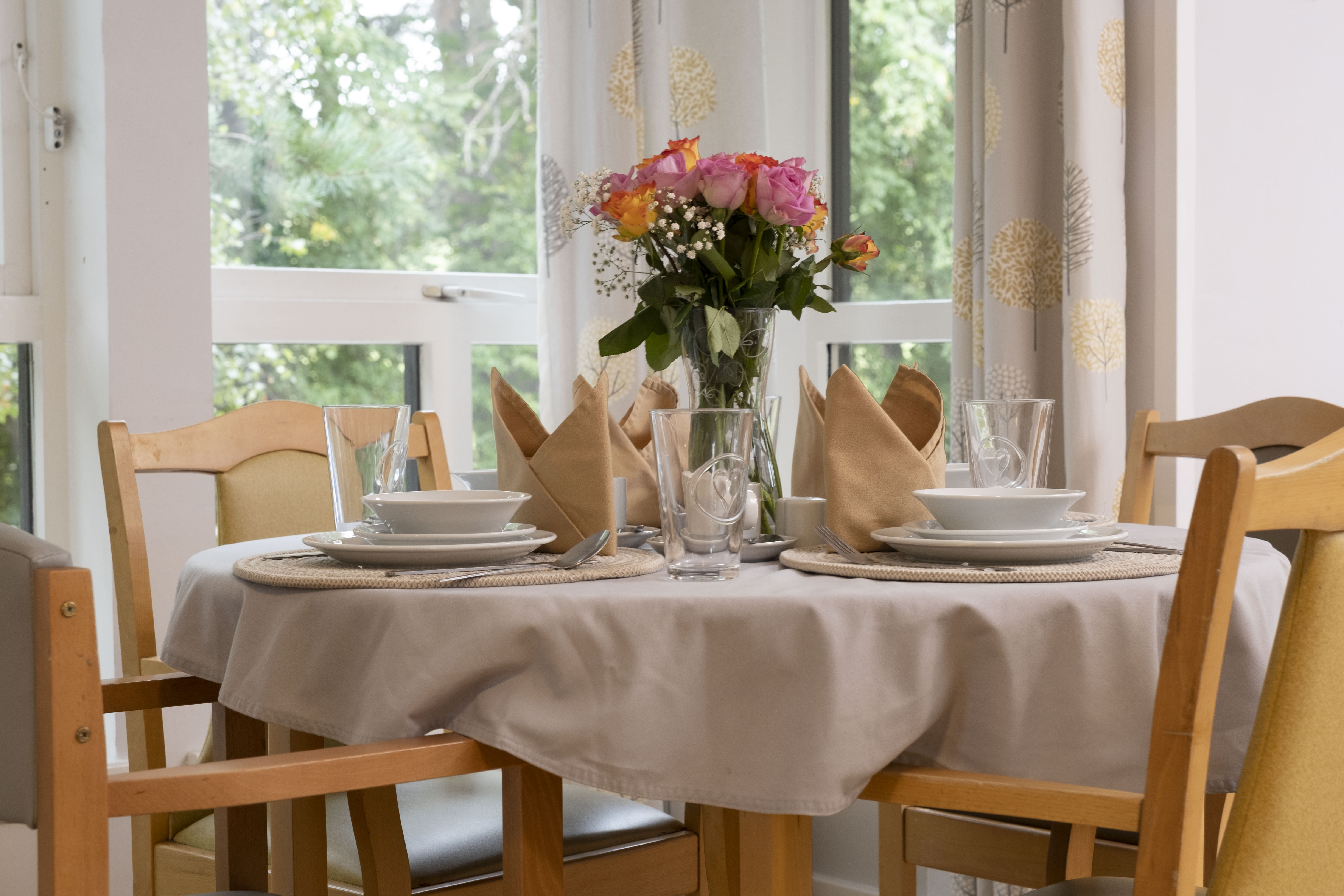 A dining room set up with a table and 4 chairs, napkins, plates, cutlery and fresh flowers. There are windows with curtains to the rear 