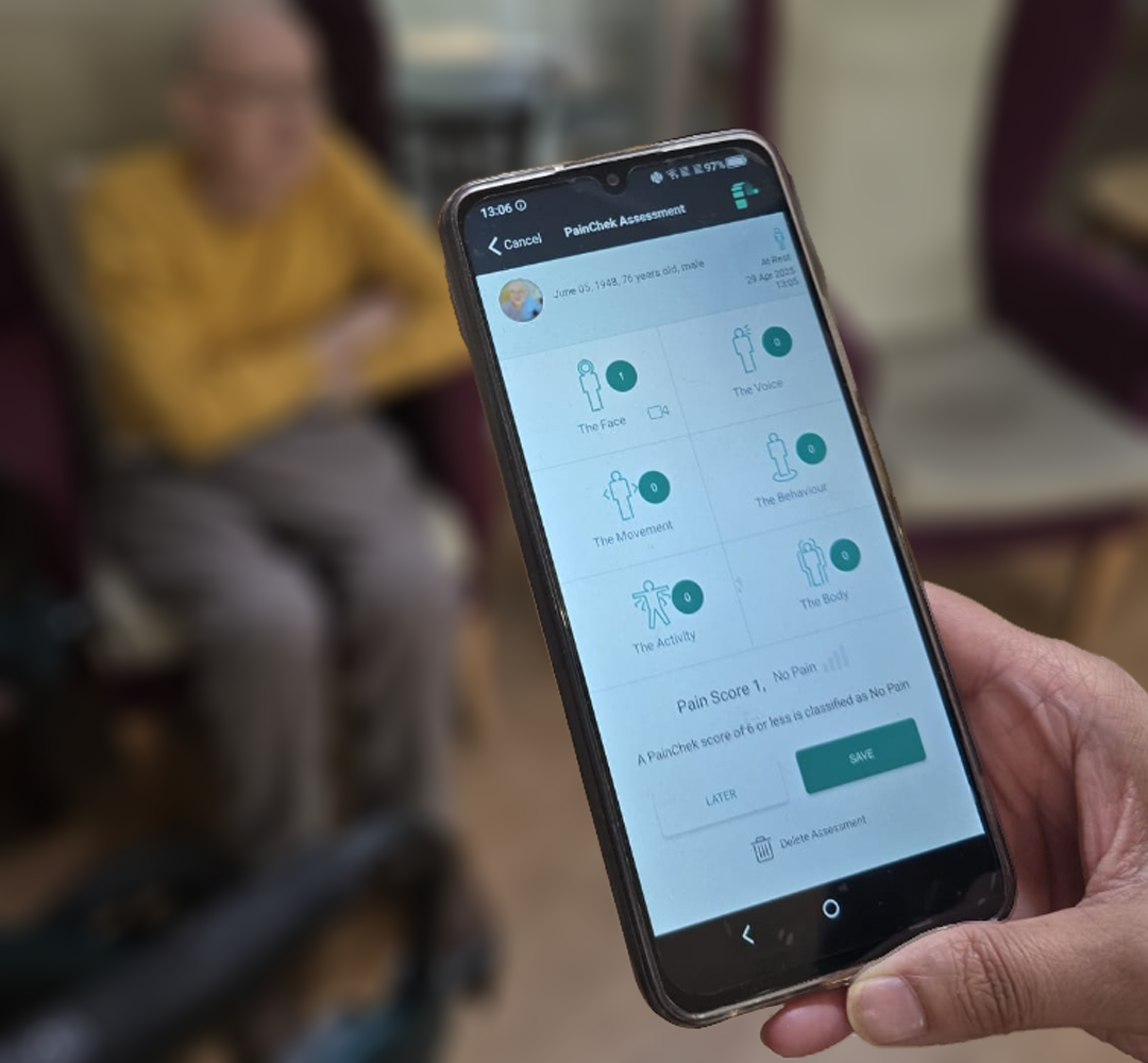 A close up shot of a phone showing a PainChek pain assessment screen. The resident who's assessment is being completed sits comfortably on a high wingbacked armchair in the background.
