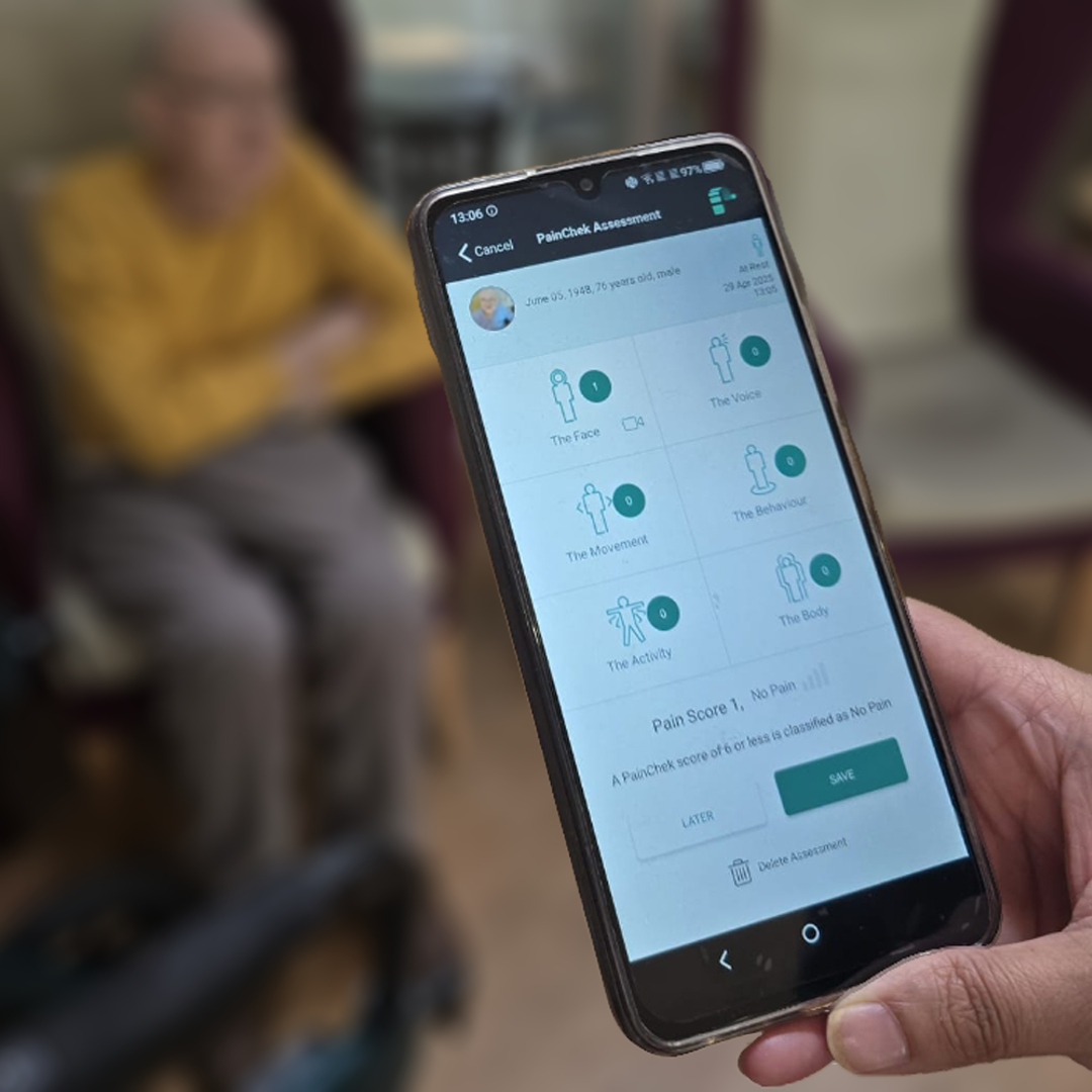 A close up shot of a phone showing a PainChek pain assessment screen. The resident who's assessment is being completed sits comfortably on a high wingbacked armchair in the background.