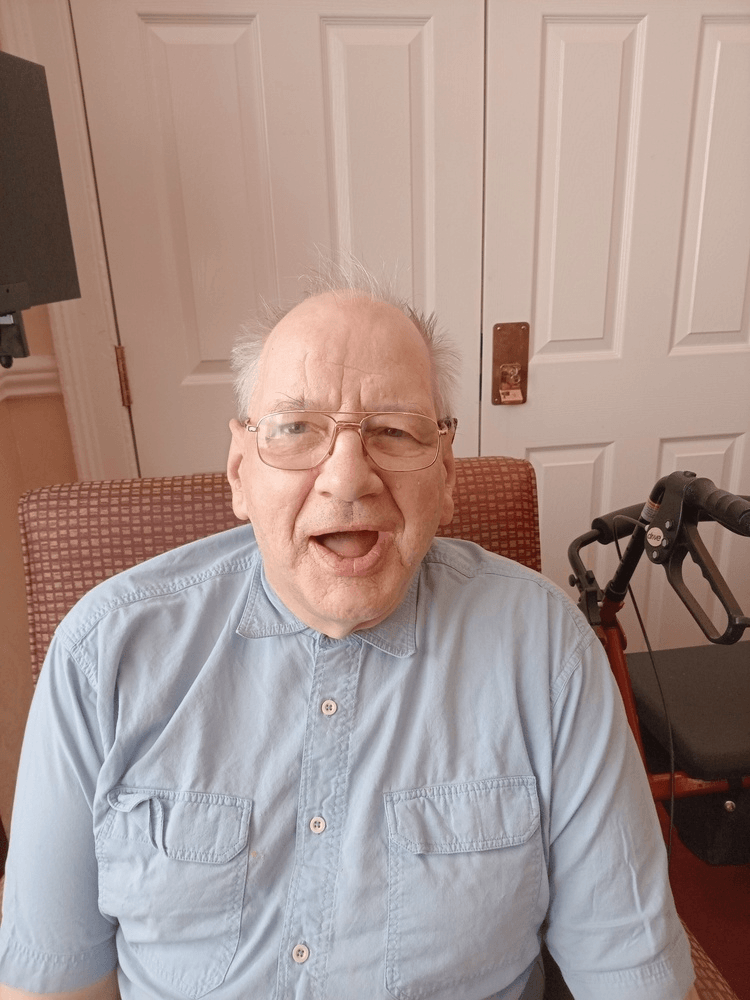 Care home resident James has a blue shirt on and glasses. He sits on a red chair.