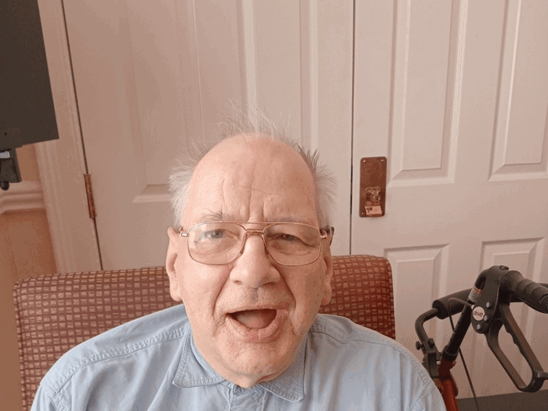 Care home resident James has a blue shirt on and glasses. He sits on a red chair.