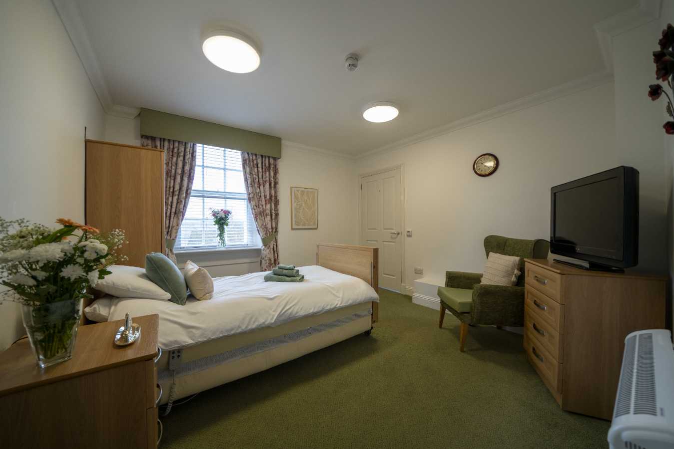 A tranquil care home bedroom with inviting sage green accents, featuring a comfortable bed, bedside unit, TV unit, and chair.