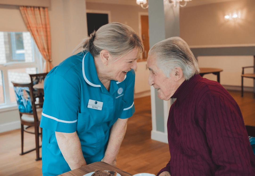A staff member in a teal uniform leans in to talk with a male resident seated at a table. They are caught in a moment of shared laughter and eye contact. In the background, the dining area is visible with floral-patterned chairs and warm lighting.