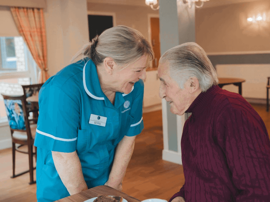 A staff member in a teal uniform leans in to talk with a male resident seated at a table. They are caught in a moment of shared laughter and eye contact. In the background, the dining area is visible with floral-patterned chairs and warm lighting.