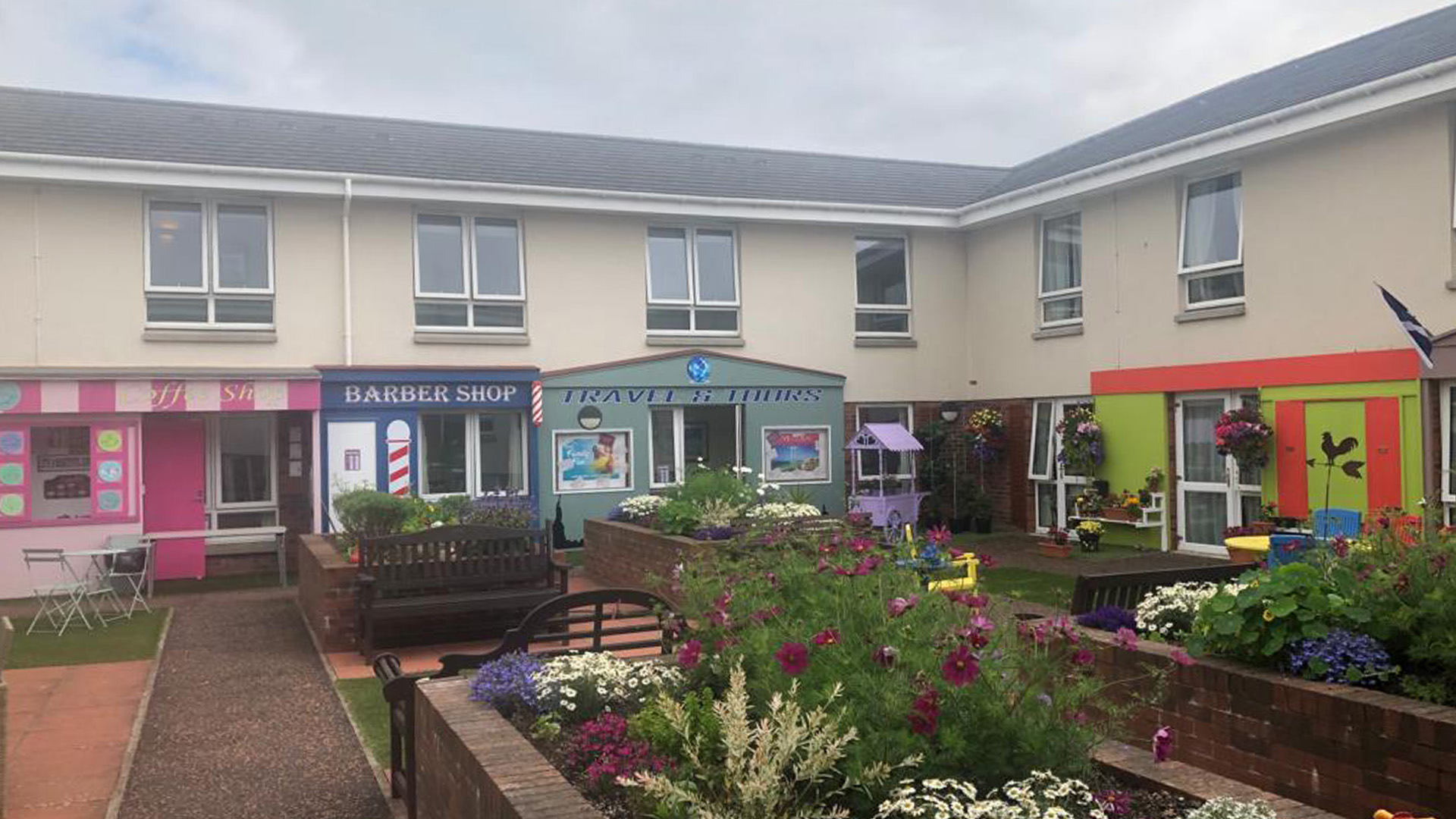 Exterior courtyard featuring shop fronts such as coffee shop and barbers. There are lots of bright colourful flowers present and a walkway