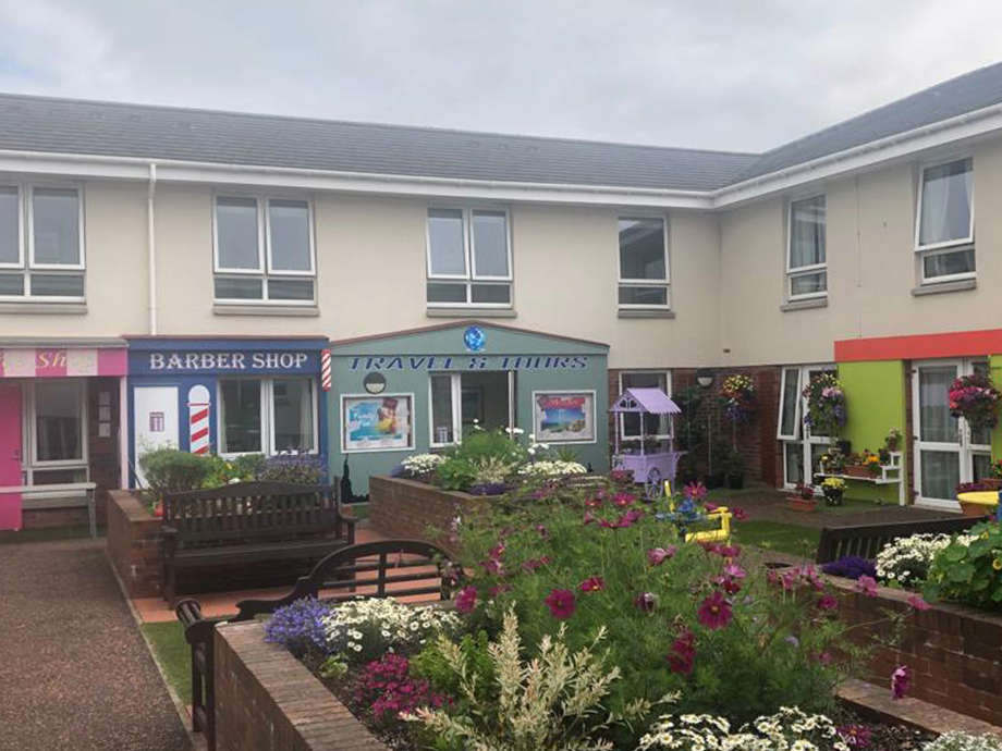 Exterior courtyard featuring shop fronts such as coffee shop and barbers. There are lots of bright colourful flowers present and a walkway