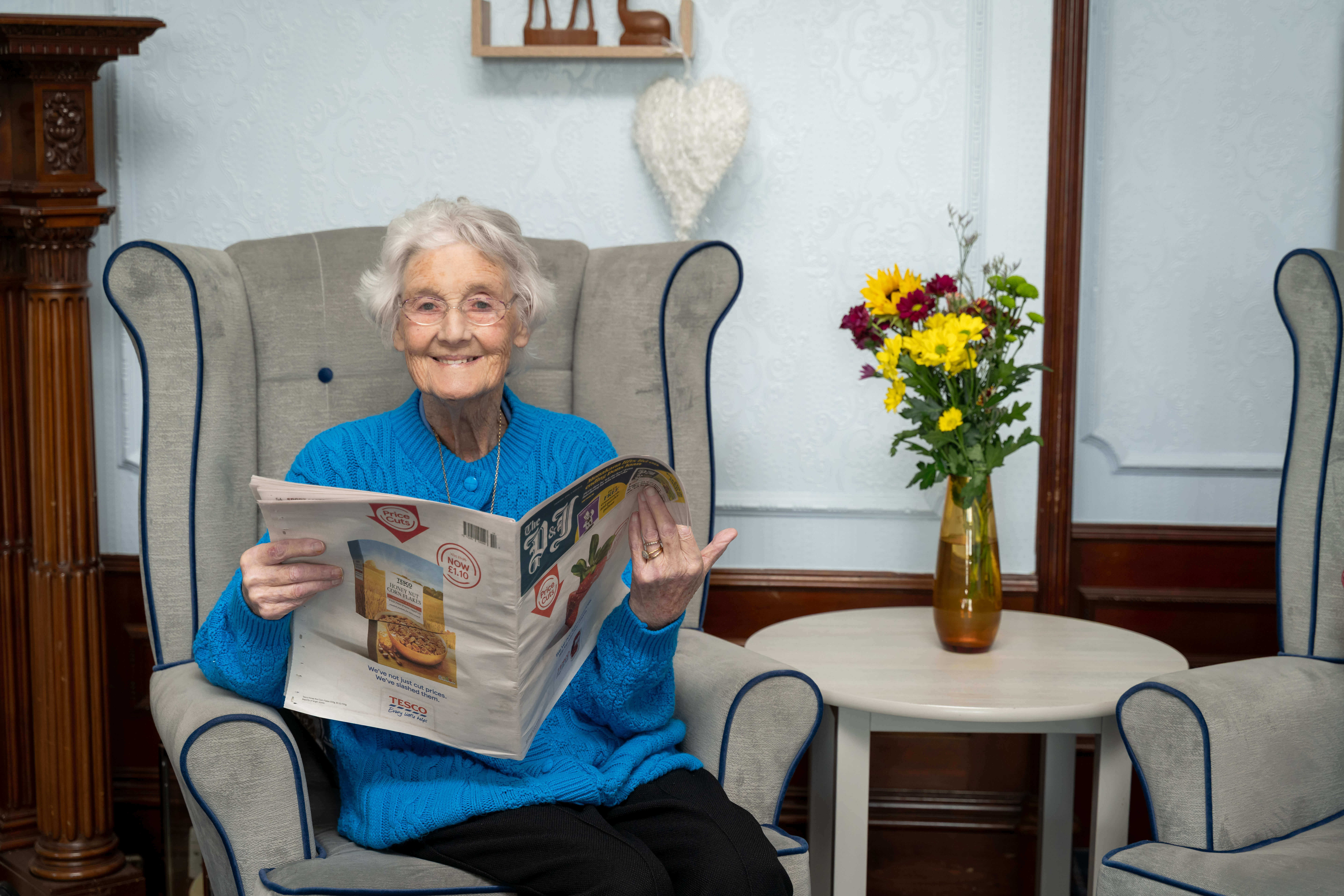 A woman in a bright blue cardigan smiles at the camera while reading a newspaper in the communal lounge, seated on a high-backed armchair.