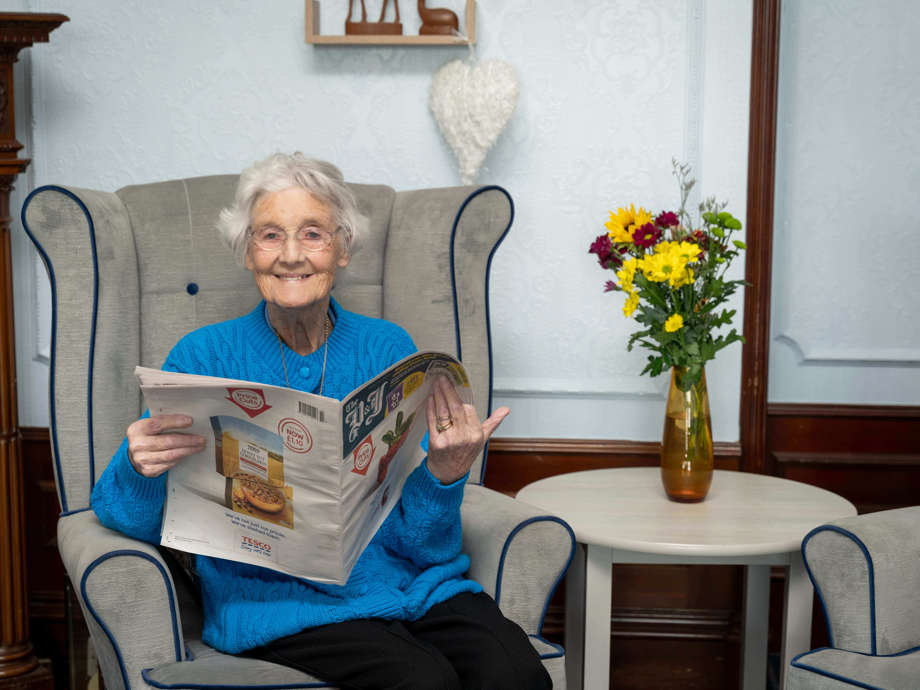 A woman in a bright blue cardigan smiles at the camera while reading a newspaper in the communal lounge, seated on a high-backed armchair.