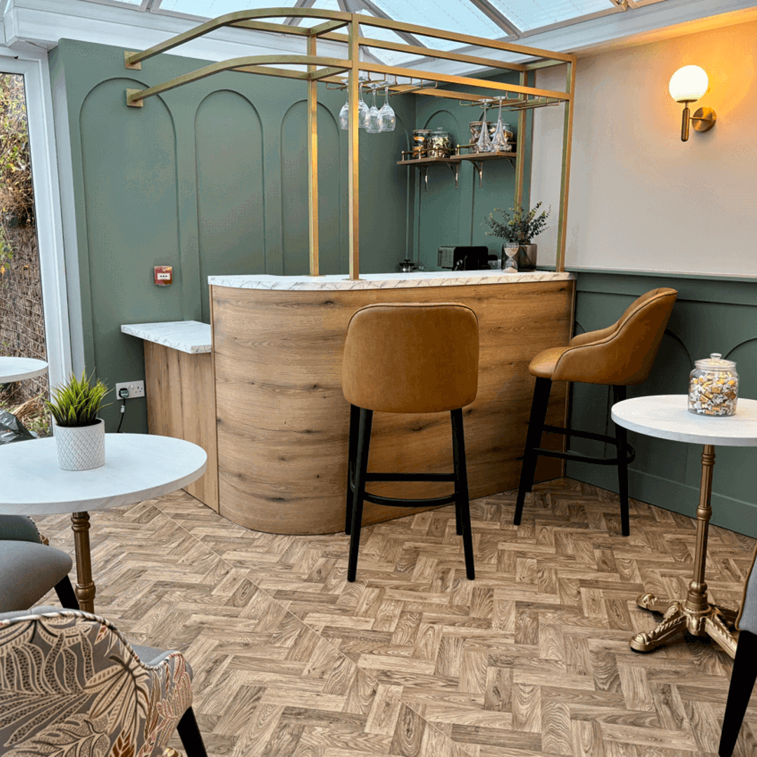 An indoor bar area in a conservatory setting. It features a curved wooden bar with a gold-toned metal frame overhead, two brown leather bar stools, and several small tables with white tops and grey upholstered chairs. The walls are painted a sage green.