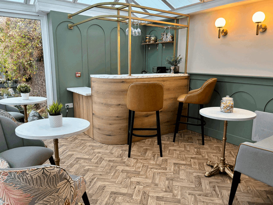 An indoor bar area in a conservatory setting. It features a curved wooden bar with a gold-toned metal frame overhead, two brown leather bar stools, and several small tables with white tops and grey upholstered chairs. The walls are painted a sage green.