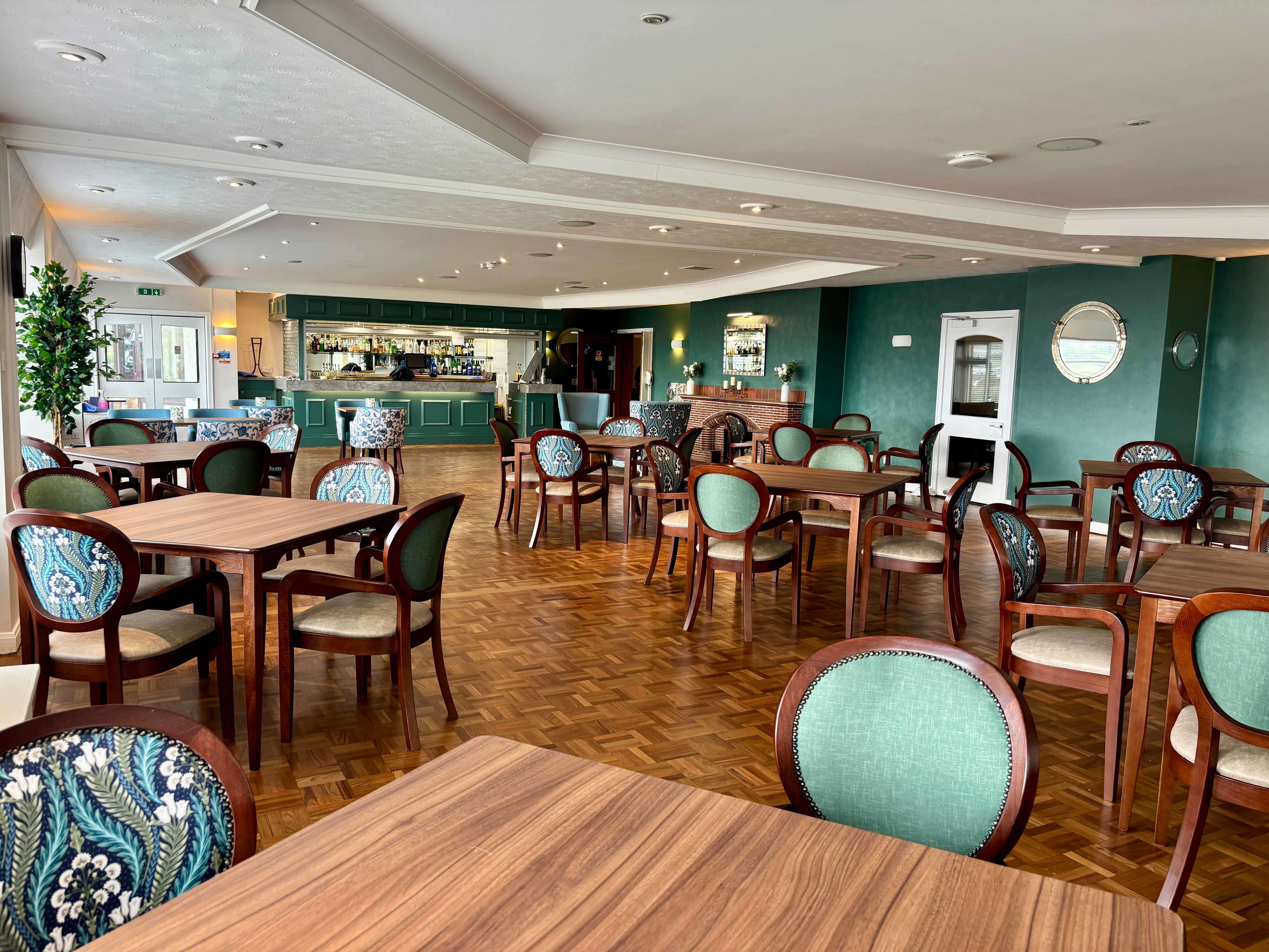 A spacious, elegant dining room with many wooden tables and chairs. The walls are a deep teal/hunter green, and a bar area with stools is visible in the background. Chairs are upholstered in a mix of solid sage green and a green/blue floral pattern.