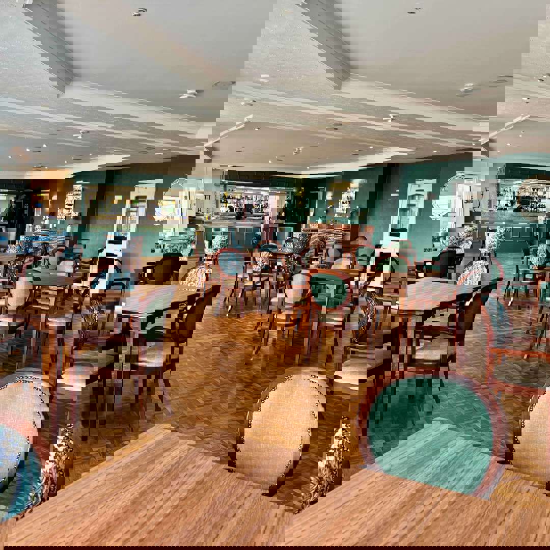 A spacious, elegant dining room with many wooden tables and chairs. The walls are a deep teal/hunter green, and a bar area with stools is visible in the background. Chairs are upholstered in a mix of solid sage green and a green/blue floral pattern.