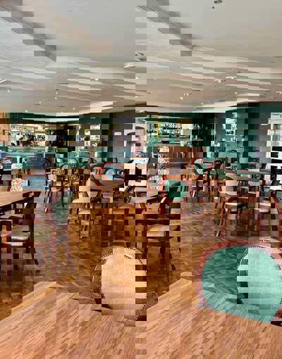 A spacious, elegant dining room with many wooden tables and chairs. The walls are a deep teal/hunter green, and a bar area with stools is visible in the background. Chairs are upholstered in a mix of solid sage green and a green/blue floral pattern.