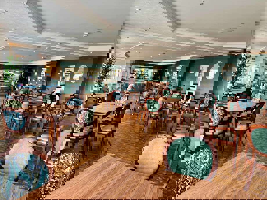 A spacious, elegant dining room with many wooden tables and chairs. The walls are a deep teal/hunter green, and a bar area with stools is visible in the background. Chairs are upholstered in a mix of solid sage green and a green/blue floral pattern.