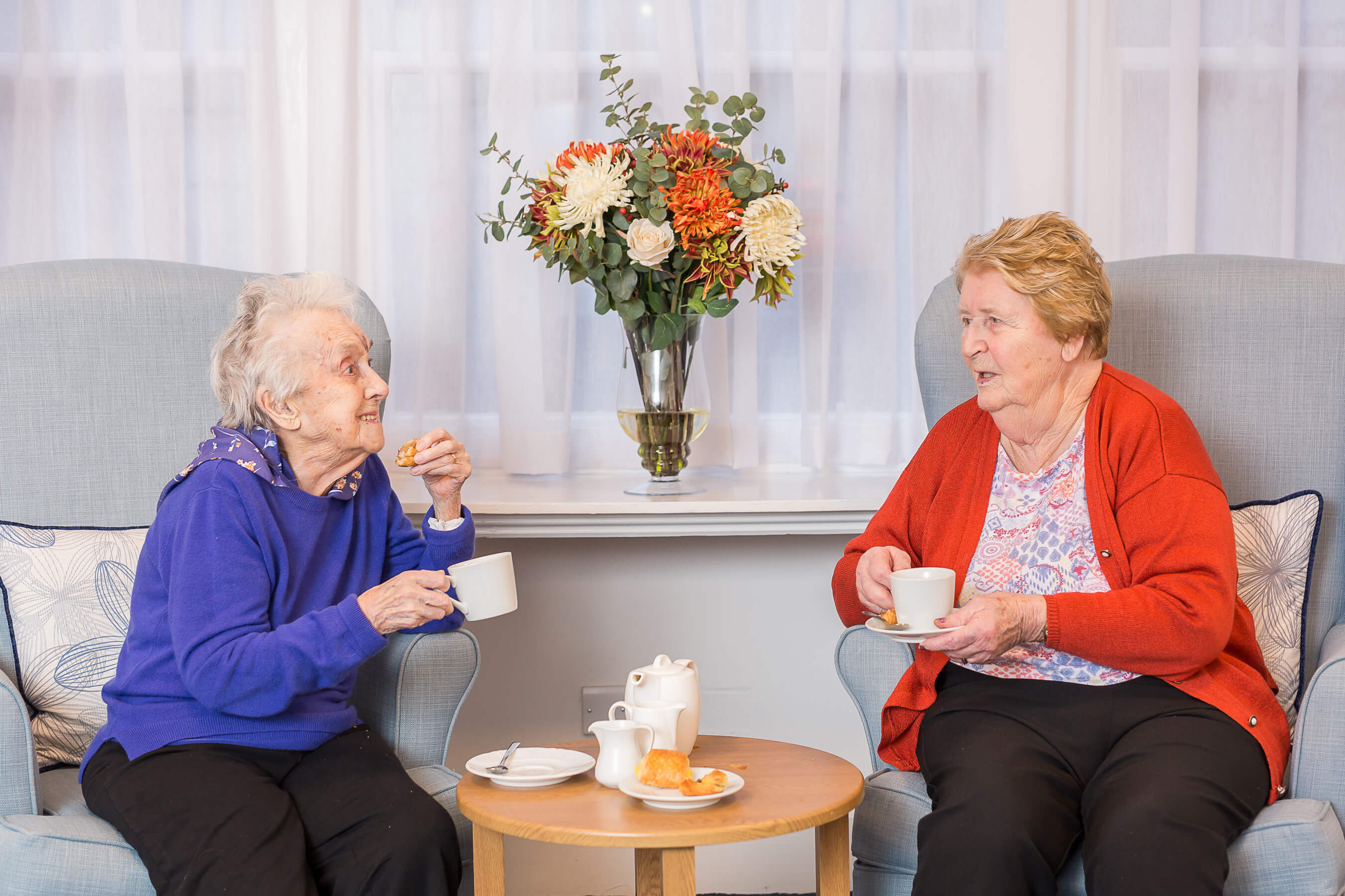 Two residents sit on high backed armchairs enjoying a cup of tea together and a chat