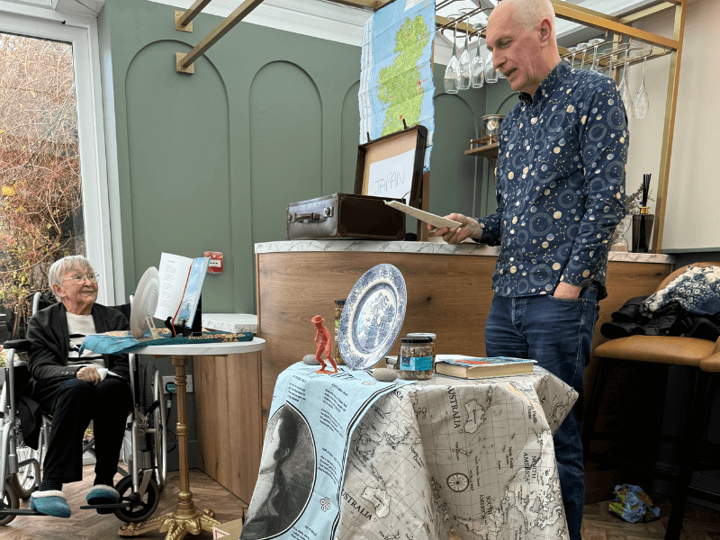 Local poet Ken Cockburn presenting to a resident during a 'paper trails' event in Glencairns conservatory area, with a table set up with travel-themed props.