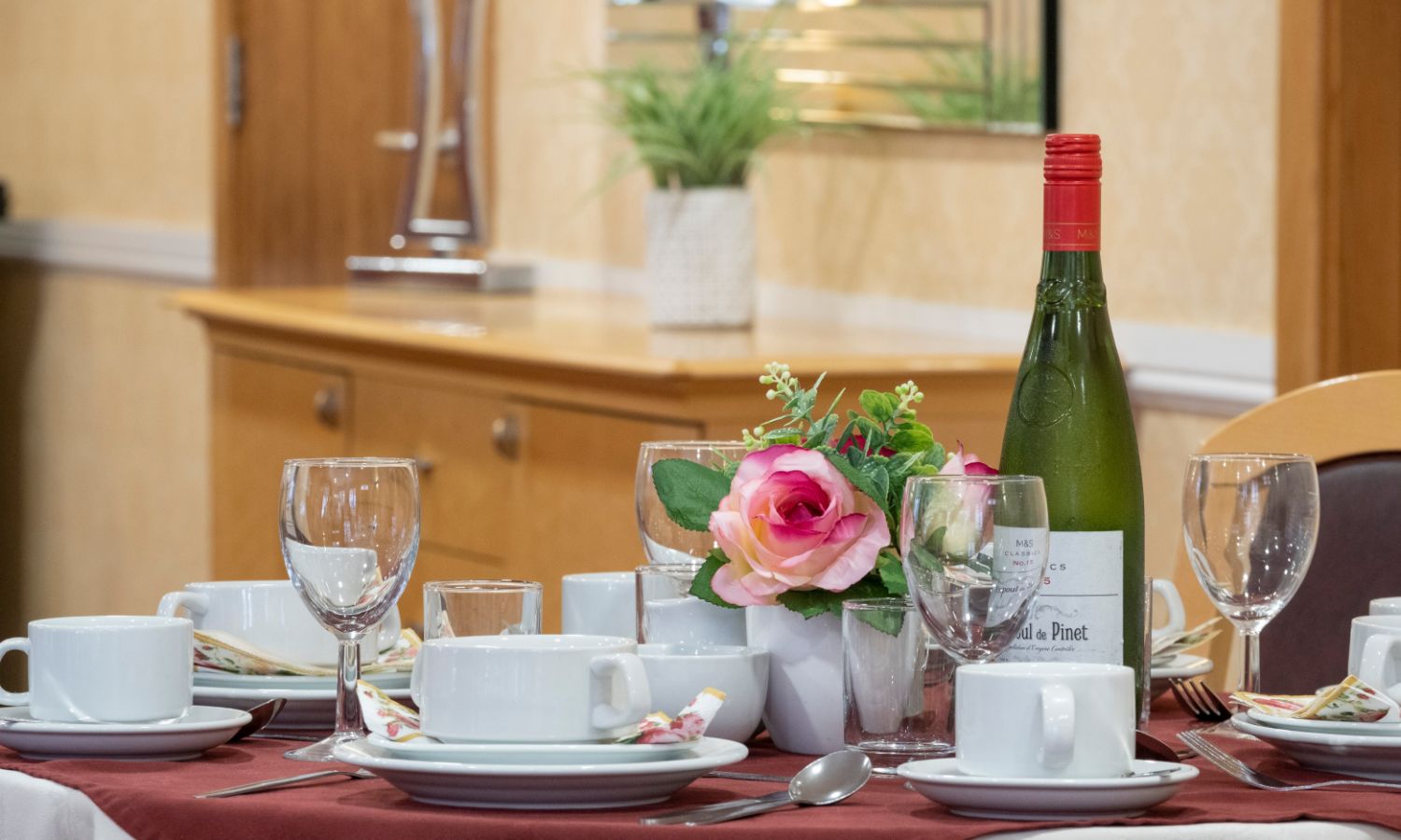 A decorative table set up in the dining room. A table with a red tablecloth and an array of teacups, wine glasses, flowers & a bottle of wine