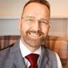 A headshot of manager Mike Culley. He has short brown hair, a beard, and glasses, and is wearing a gray waistcoat over a white shirt and a red tie. The background is slightly blurred and features a light-coloured shelf and a patterned armchair.