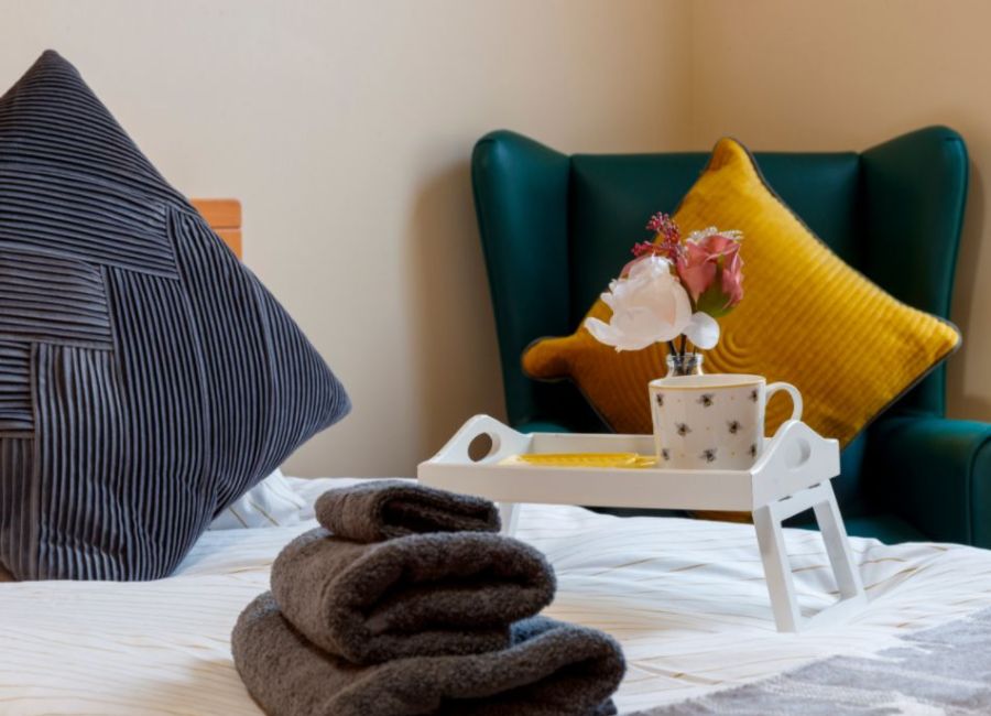 A bedroom set up with a white duvet, fluffy towels, a breakfast tray, and armchair