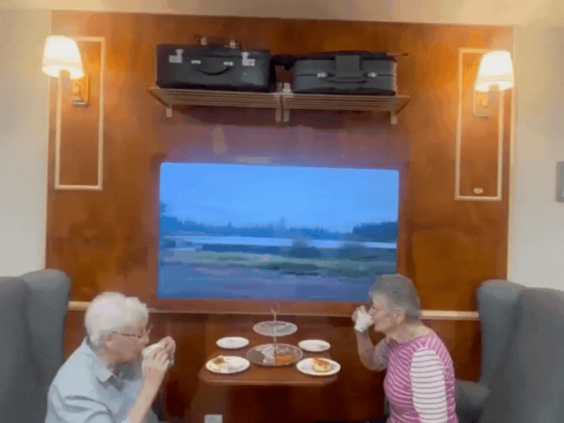 Two residents sit and have high tea in the home at an area set up like a train carriage - suitcases sit overhead with a tv displaying a train scene view on the wall