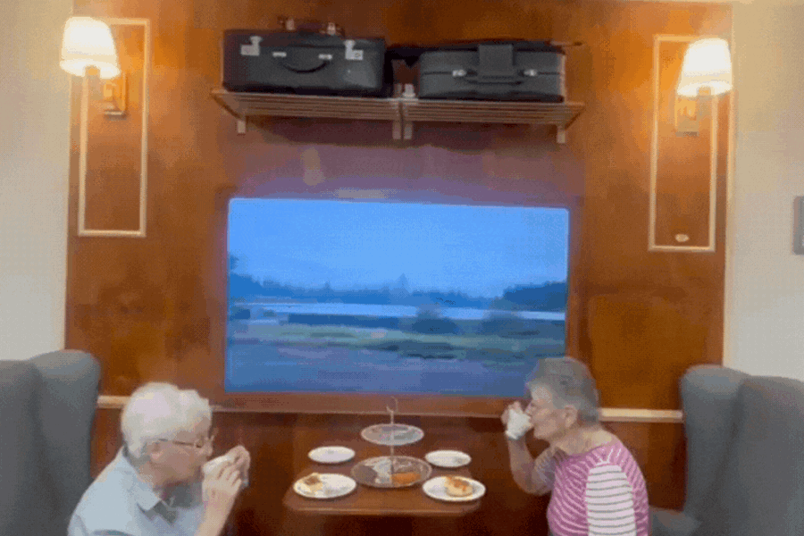 Two residents sit and have high tea in the home at an area set up like a train carriage - suitcases sit overhead with a tv displaying a train scene view on the wall