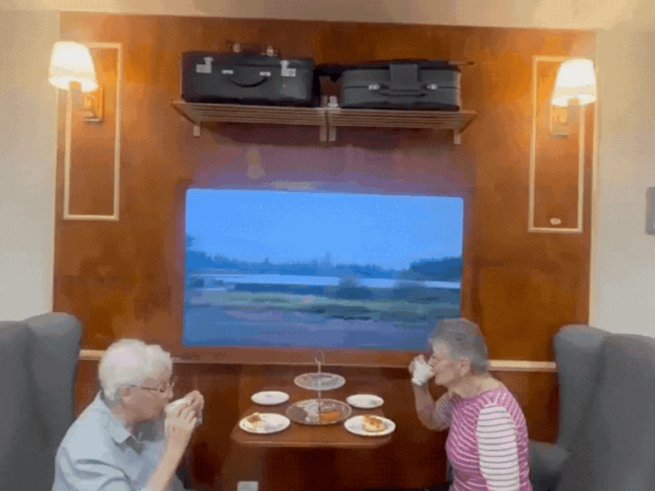 Two residents sit and have high tea in the home at an area set up like a train carriage - suitcases sit overhead with a tv displaying a train scene view on the wall