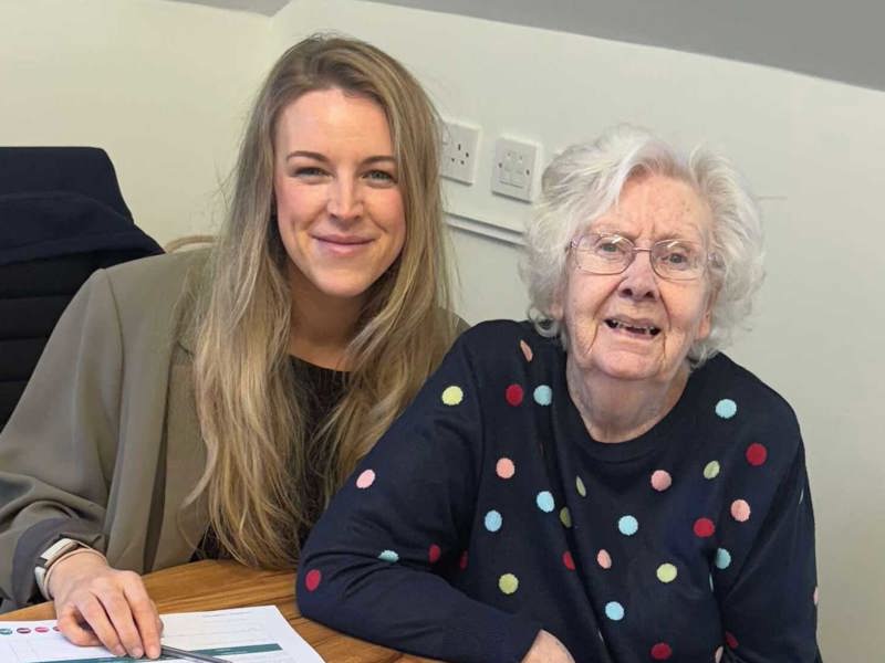 Resident Mairi wears a spotted navy jumper and sits at a wooden table with Home Manager Jozi Stables